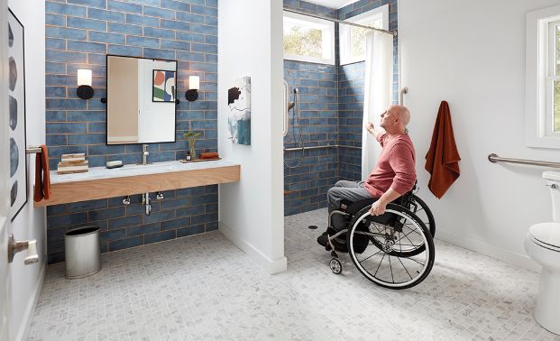 A man  in a wheelchair in an accessible bathroom with grab bars and a zero entry shower. The bathroom sink is open underneath allowing a wheelchair user to reach the sink comfortably.