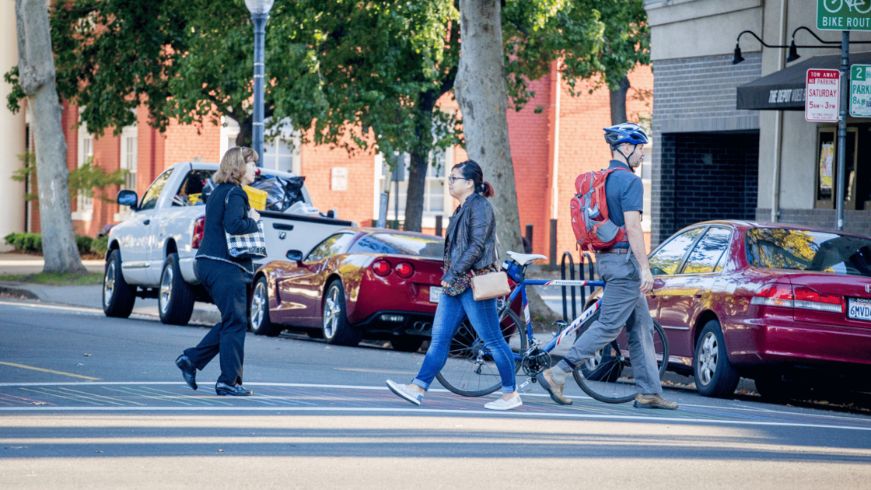 Three people using a crosswalk, cars parked, bicycle