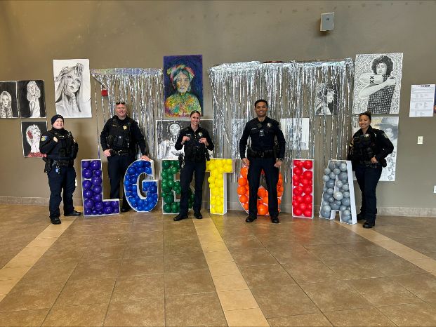 A photo of 5 Sacramento Police Department LGBTQ+ liaison officers, in full uniform, standing intermixed among the letters L G B T Q I  and A which are each painted a different color of the rainbow. On the wall behind them are various works of art from our local LGBTQIA+ community.