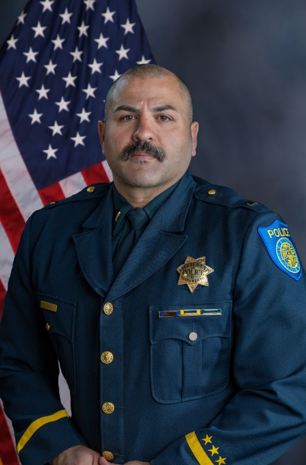 Photo of Sameer Sood in uniform standing in front of the American flag.