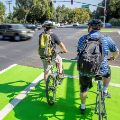 Photo of two bicyclists waiting in a green two-stage left turn box to cross a busy intersection.